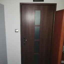 Ciemnobrązowe drzwi wewnętrzne z trzema pionowymi szybkami, zamontowane w białej ścianie, nad drzwiami wisi obrazek religijny w czarnej ramce.