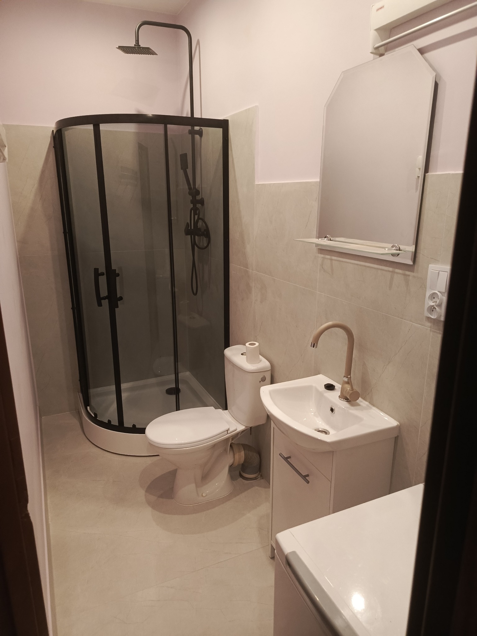 Nowoczesna łazienka po remoncie: kabina prysznicowa z czarnymi profilami, biała umywalka z szafką i lustro na tle beżowych płytek. Minimalistyczny design.