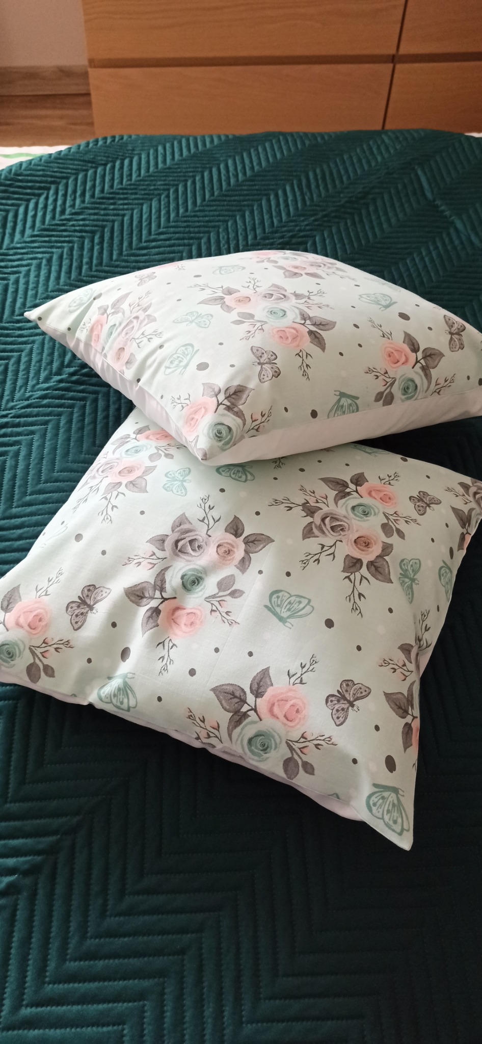 Dwa kwadratowe jaśki z delikatnym wzorem róż i motyli leżące na ciemnozielonej, pikowanej narzucie na łóżko.