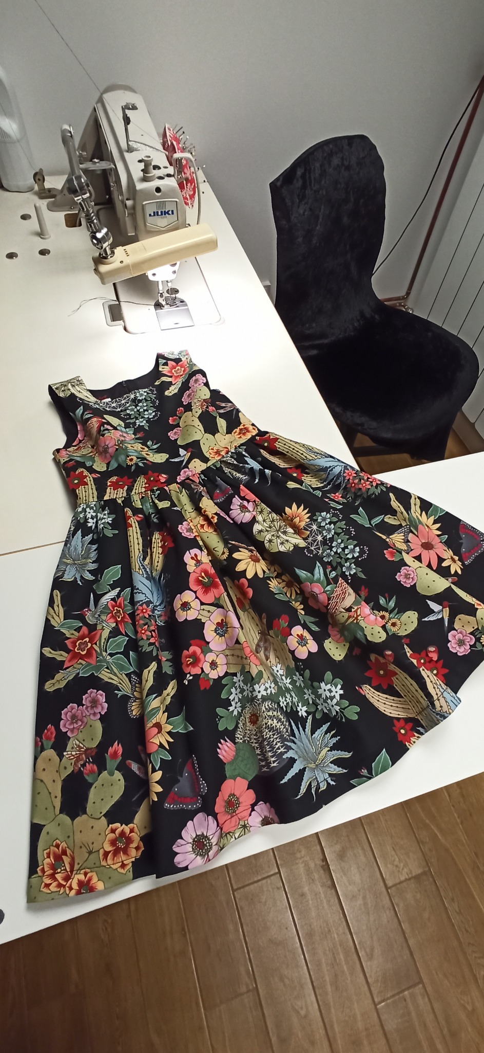 Gotowa sukienka z czarnej tkaniny w motywy kaktusów i kwiatów leży na białym stole obok maszyny do szycia JUKI i czarnego krzesła.