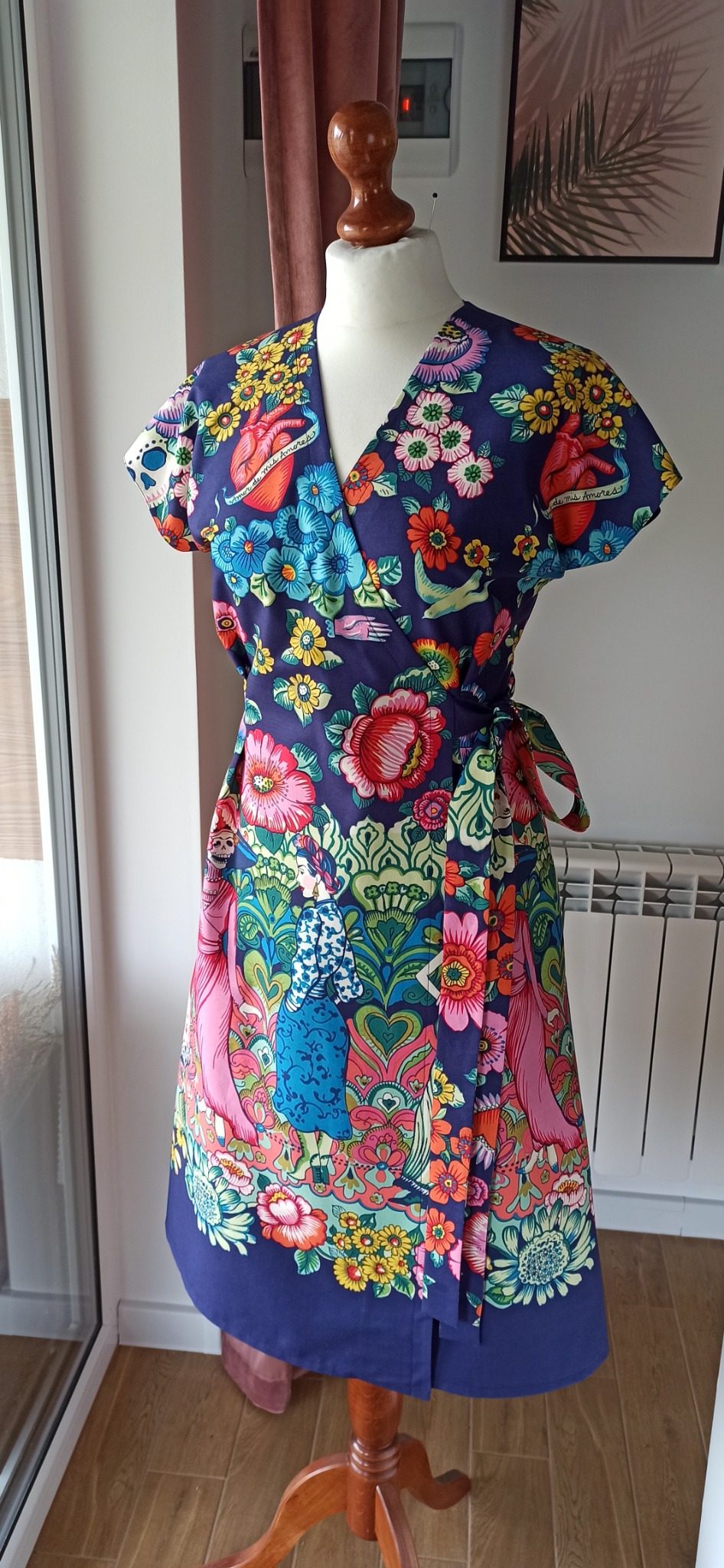 Sukienka z motywem kwiatowym i meksykańskimi elementami, zapinana na zakładkę, prezentowana na manekinie krawieckim w jasnym pomieszczeniu.