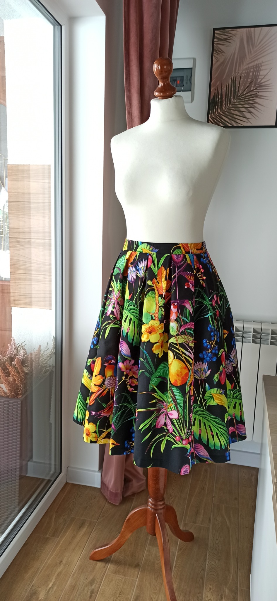 Rozkloszowana spódnica z czarnego materiału w tropikalne wzory, prezentowana na białym manekinie krawieckim na drewnianej podstawie, w jasnym wnętrzu z oknem i zasłoną.