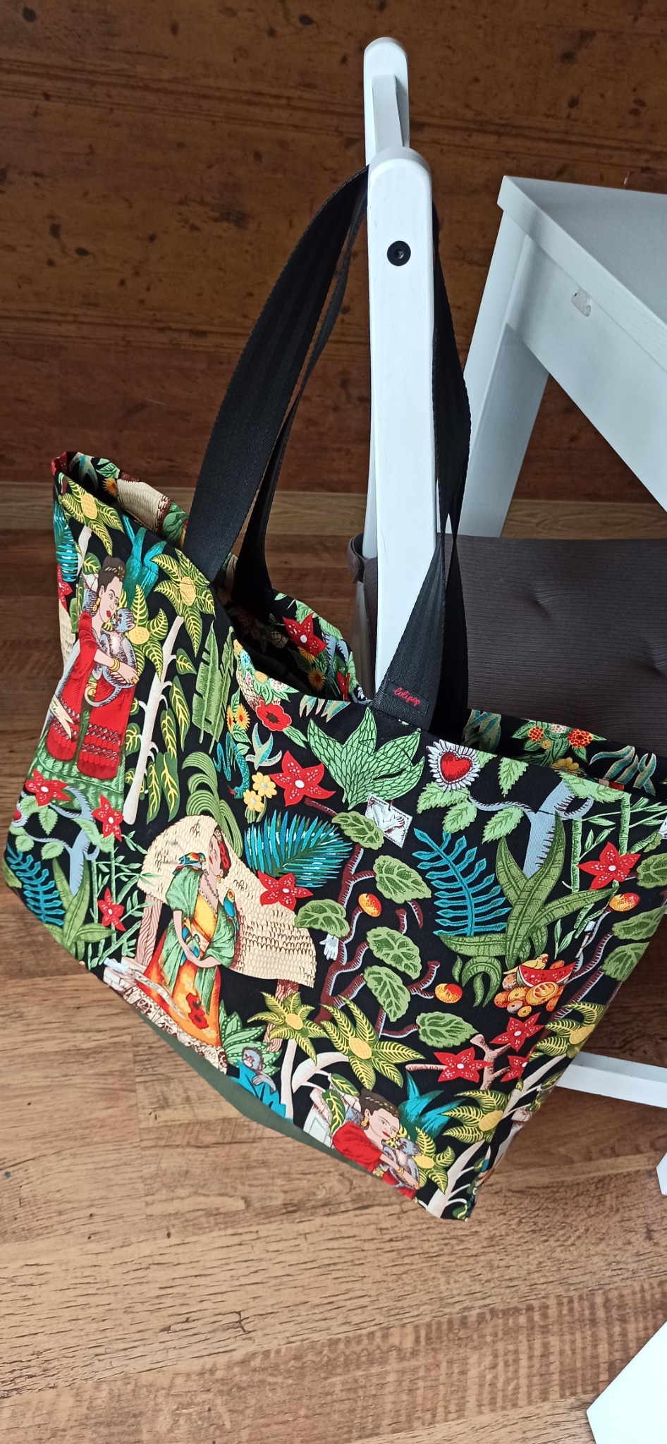 Torba shopper z czarnymi uchwytami, wisząca na białym krześle, z wzorem inspirowanym dżunglą i postaciami kobiecymi w stylu vintage.