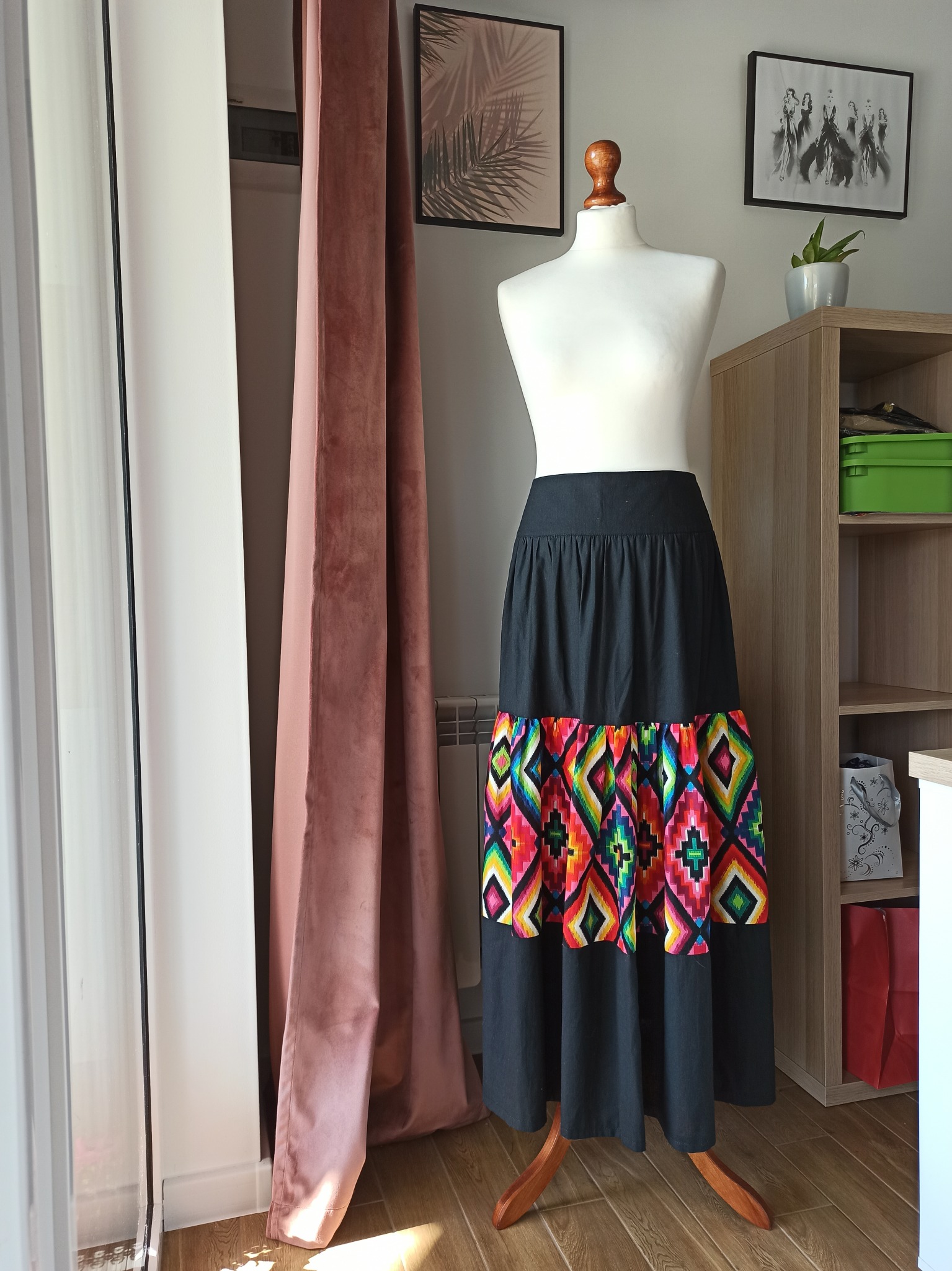 Długa czarna spódnica z kolorowym, etnicznym wzorem na manekinie krawieckim, ustawionym w jasnym pomieszczeniu obok okna z zasłoną.