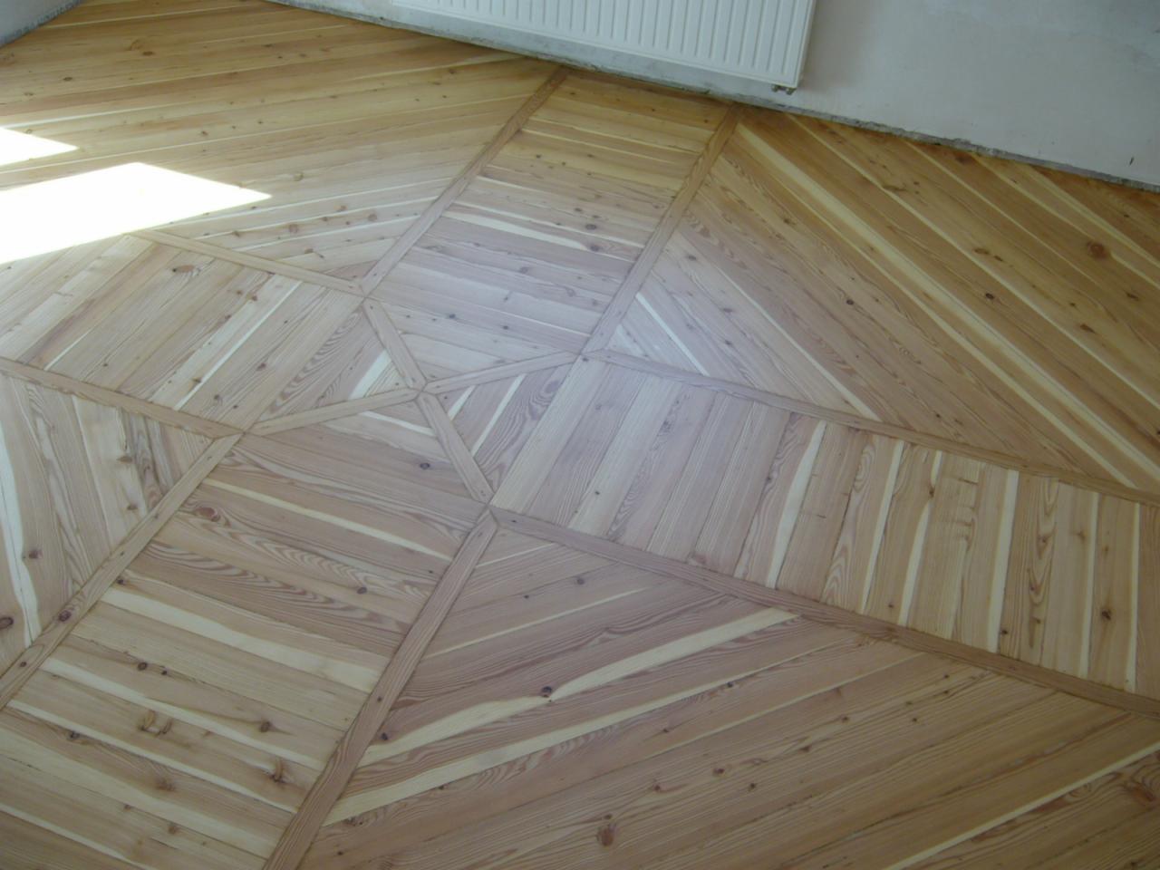 Ułożona podłoga parkietowa z jasnego drewna w geometryczny wzór z widocznymi liniami łączeń i fragmentem grzejnika w rogu.