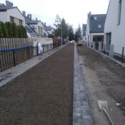 Przygotowanie terenu pod ścieżkę między domami: wyrównana ziemia, obrzeża z kostki, widoczny mini-koparka i łopata, sugerujące prace ziemne.
