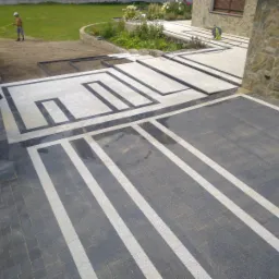 Ułożona kostka brukowa w dwóch odcieniach szarości z białymi liniami, tworząca geometryczny wzór na podjeździe, częściowo mokra powierzchnia po czyszczeniu, w tle trawnik i fragment domu z kamienną...