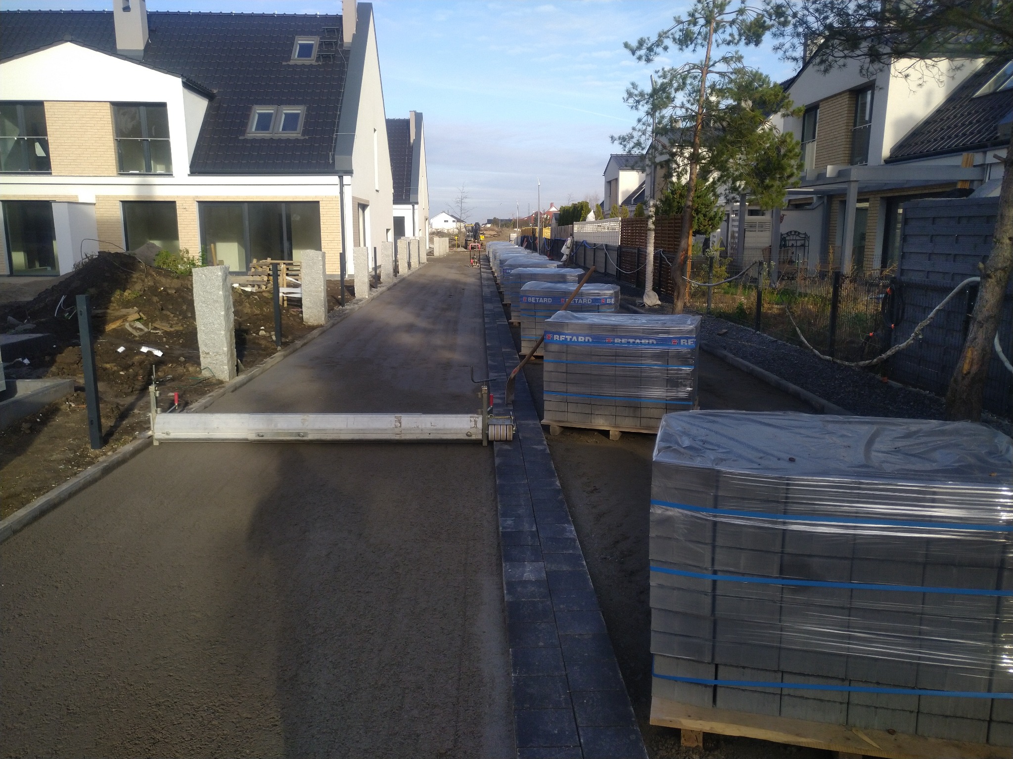 Przygotowanie podłoża pod kostkę brukową na osiedlowej drodze, widoczne palety z kostką, listwa wyrównująca i obrzeża chodnikowe.