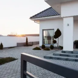 Nowoczesny dom jednorodzinny z podjazdem z szarej kostki brukowej, niskie krzewy ozdobne. W tle biały mur i zachodzące słońce.