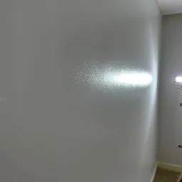 Świeżo pomalowana szara ściana w rogu pokoju, z widocznym punktowym oświetleniem halogenowym uwydatniającym fakturę i połysk farby, zabezpieczona taśmą malarską przy podłodze i suficie.