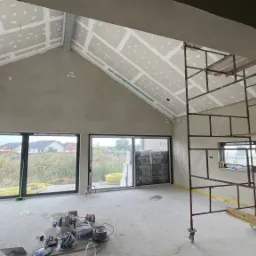 Wnętrze w stanie deweloperskim z zamontowanymi płytami kartonowo-gipsowymi na skośnym suficie, widoczne szpachlowane ściany, rusztowanie i narzędzia na podłodze.