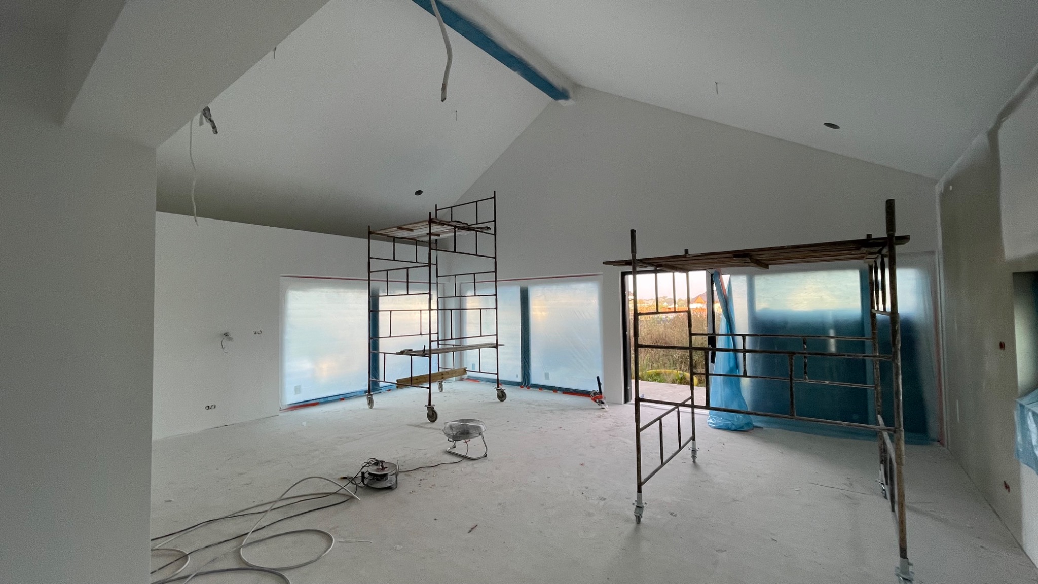 Wnętrze w trakcie prac przygotowawczych, zabezpieczone okna folią, widoczne rusztowania, wentylator i przewody na podłodze, surowy stan ścian i sufitu przed wykończeniem.