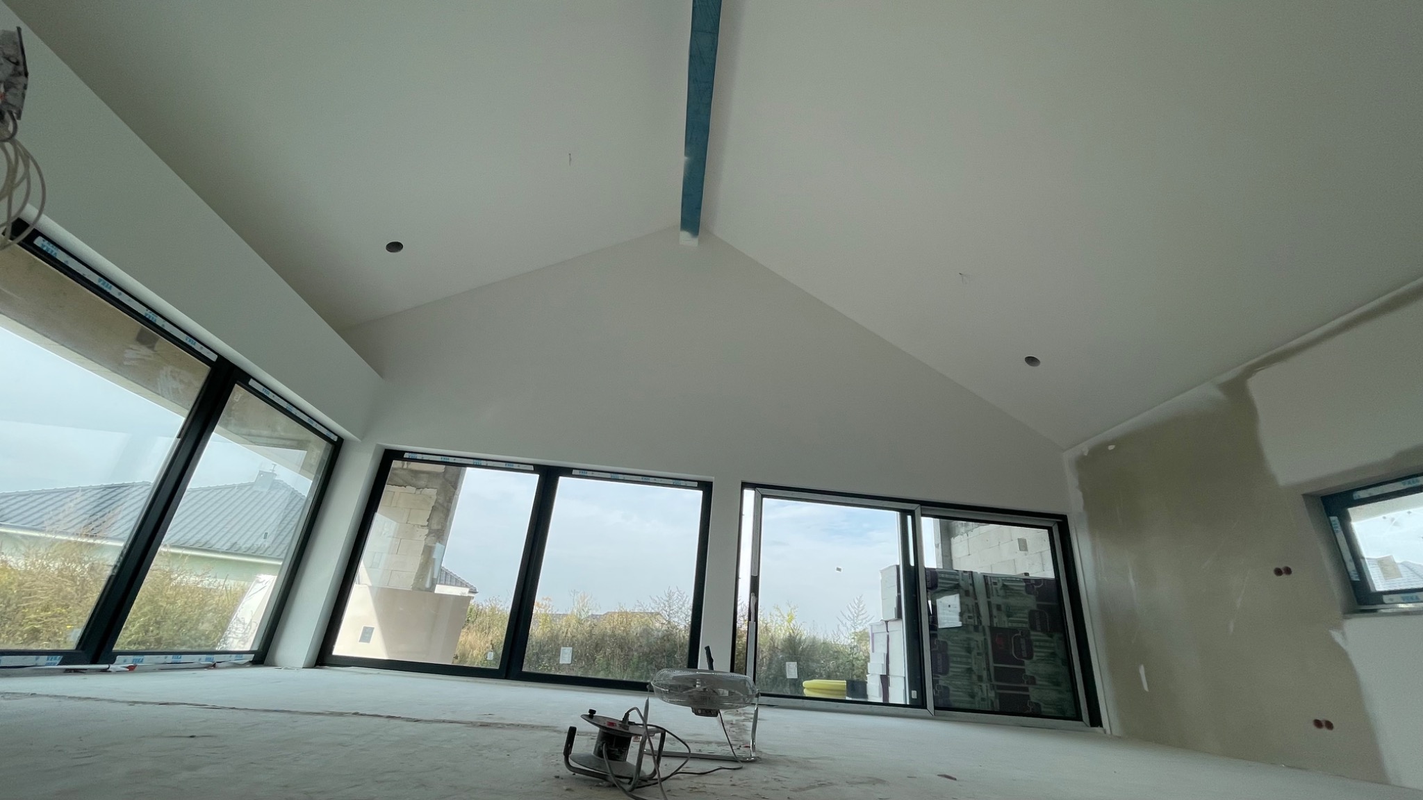 Wnętrze w stanie deweloperskim z dużymi oknami, widoczne ślady po szpachlowaniu na ścianach, wentylator podłogowy, przygotowane otwory na oświetlenie w suficie.