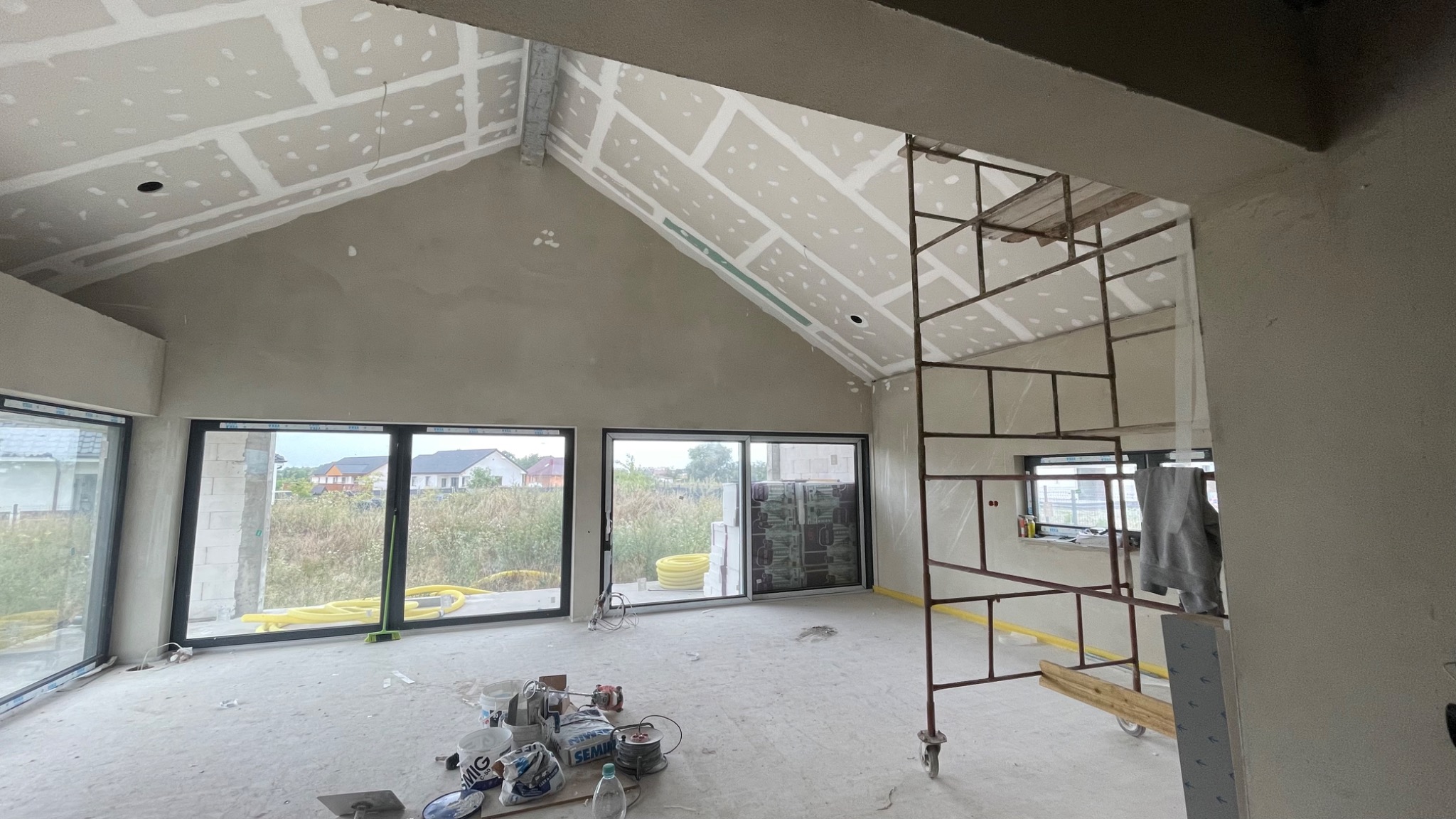 Wnętrze w stanie deweloperskim z zamontowanymi płytami kartonowo-gipsowymi na skośnym suficie, widoczne szpachlowane ściany, rusztowanie i narzędzia na podłodze.