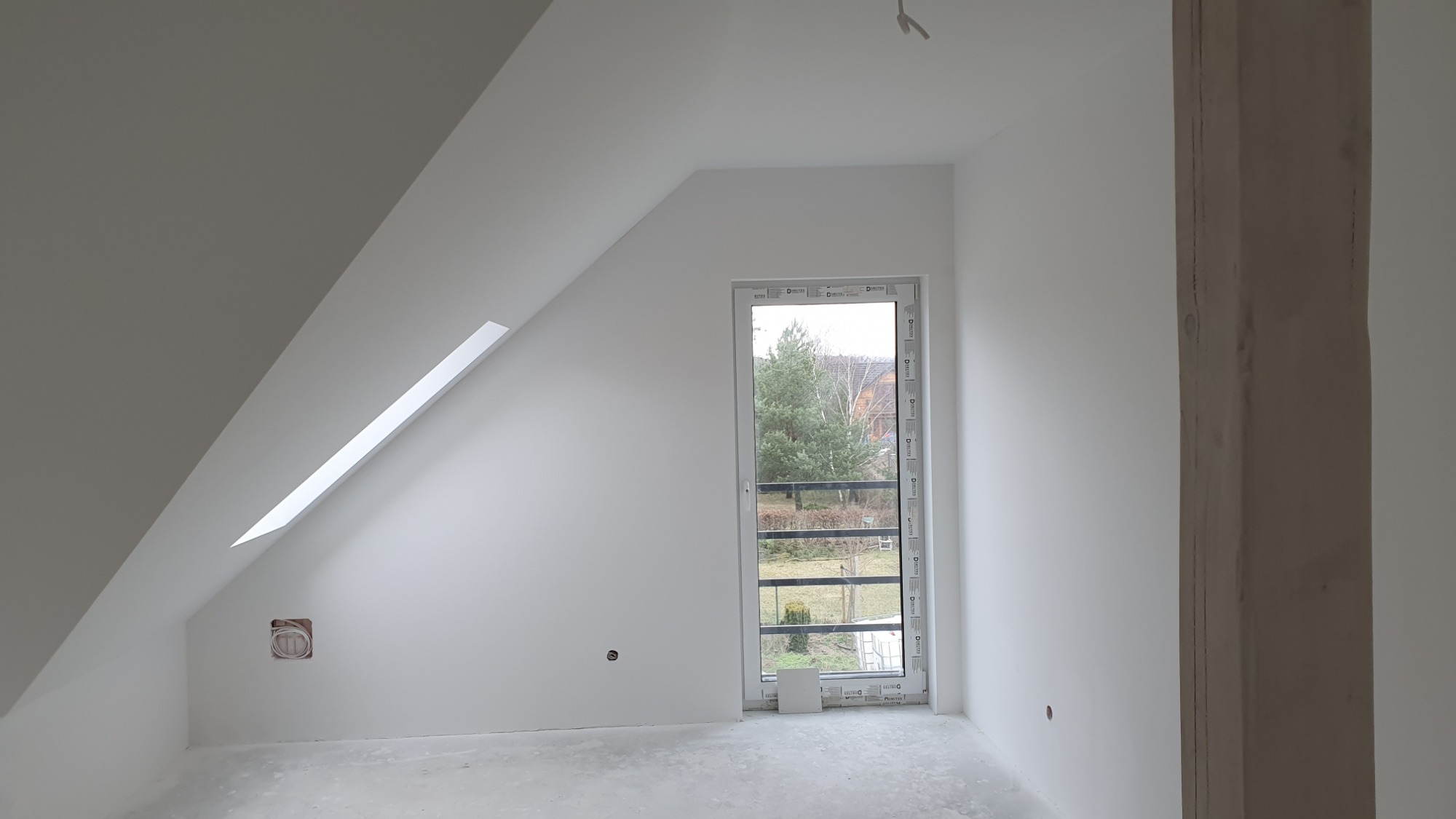 Poddasze po szpachlowaniu i malowaniu na biało, widoczne okno dachowe, okno pionowe z widokiem na zewnątrz oraz drewniany słup konstrukcyjny