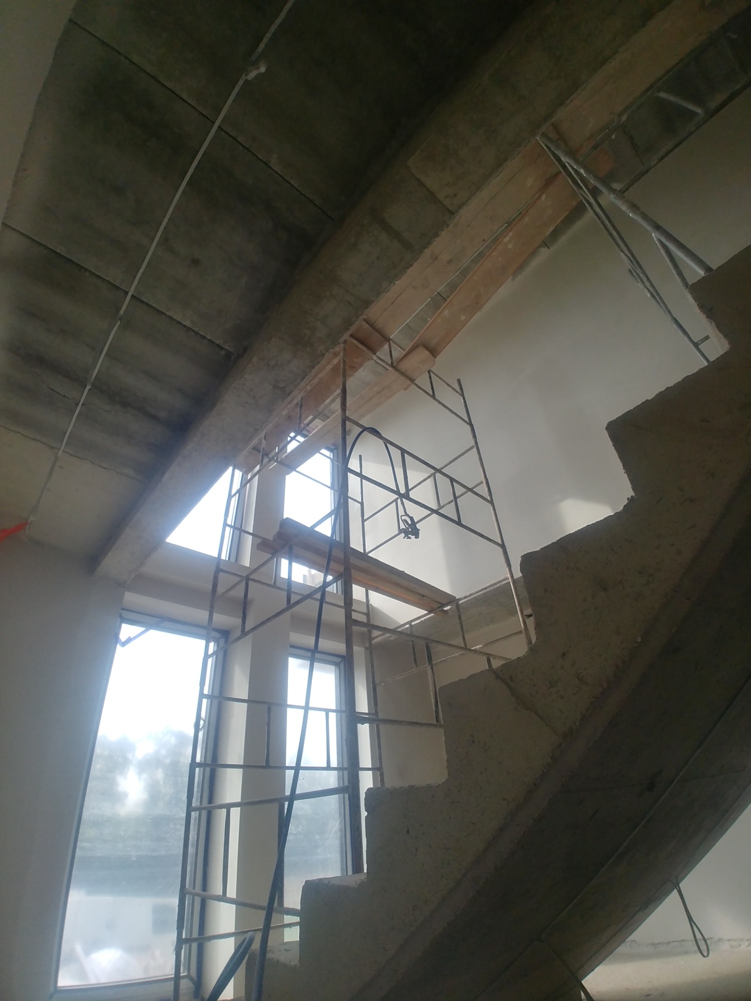 Wnętrze w trakcie prac wykończeniowych: surowy betonowy strop, fragment schodów i metalowe rusztowanie przed oknem.