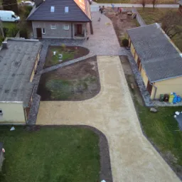 Widok z góry na posesję z nową żwirową ścieżką i podjazdem, obrzeża wykończone kostką, w tle dom jednorodzinny i budynki gospodarcze.