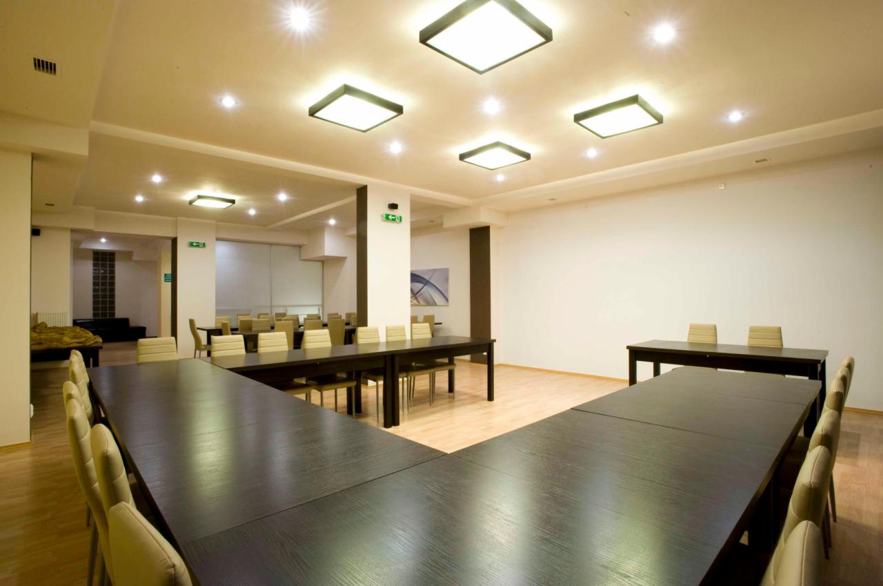 Jasne wnętrze sali szkoleniowo-bankietowej z długimi, ciemnymi stołami ustawionymi w kształt litery U, krzesłami z jasną tapicerką i kwadratowymi lampami sufitowymi.