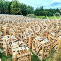 Szeroki kadr na pole pełne drewnianych skrzyń wypełnionych pociętym drewnem kominkowym, w tle las i linia energetyczna.