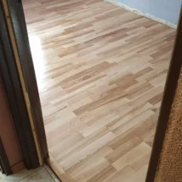 Nowo ułożona podłoga z paneli laminowanych w odcieniach jasnego drewna, widoczna z perspektywy progu, z fragmentem płytek podłogowych w sąsiednim pomieszczeniu.