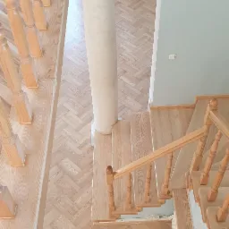 Widok z góry na drewniane schody z jasnego drewna, z kolumną dekoracyjną i parkietem ułożonym w jodełkę.