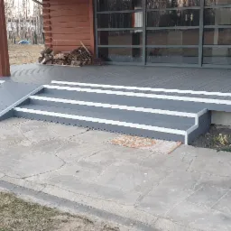 Szary taras kompozytowy z rampą dla wózków inwalidzkich, prowadzący do drewnianego domu z panoramicznymi oknami, na zewnątrz ułożona kamienna ścieżka.