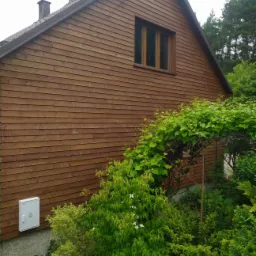 Ściana domu w Szydłowcu wykończona brązową, poziomą boazerią drewnianą, widoczna skrzynka elektryczna, okno z ciemnymi ramami i bujna zieleń ogrodu.