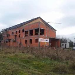 Dwukondygnacyjny, surowy budynek z czerwonej cegły w trakcie budowy, z widocznym rusztowaniem i dźwigiem w tle. Na budynku widoczny baner reklamowy.