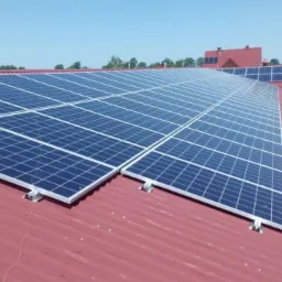 Instalacja paneli słonecznych na dachu z blachodachówki w kolorze ceglastym. Widoczne panele ułożone rzędami, mocowane do dachu. W tle budynek z żółtą ścianą i rusztowaniem oraz fragmenty innych...