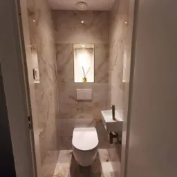 Wąska toaleta wykończona marmurowymi płytkami z podświetlaną wnęką na dekoracyjny dyfuzor zapachowy, minimalistyczną umywalką i złotą armaturą.