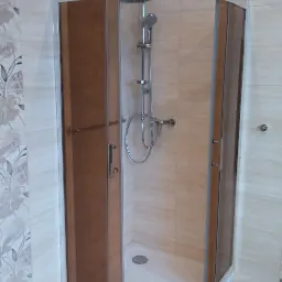 Narożna kabina prysznicowa z brązowymi drzwiami i chromowaną armaturą na tle beżowych płytek z delikatnym kwiatowym wzorem.