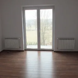 Puste, jasne pomieszczenie z brązowymi panelami podłogowymi, białymi ścianami i grzejnikami, z widokiem na zewnątrz przez okno balkonowe.