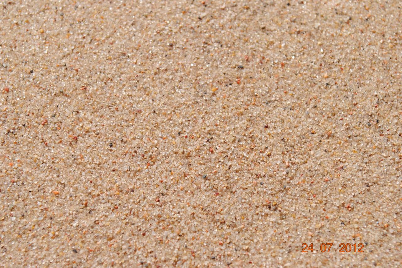 Zbliżenie na drobny, jasny piasek kwarcowy o zróżnicowanej granulacji, z widocznymi ciemniejszymi i czerwonymi ziarenkami, oświetlony jasnym światłem.