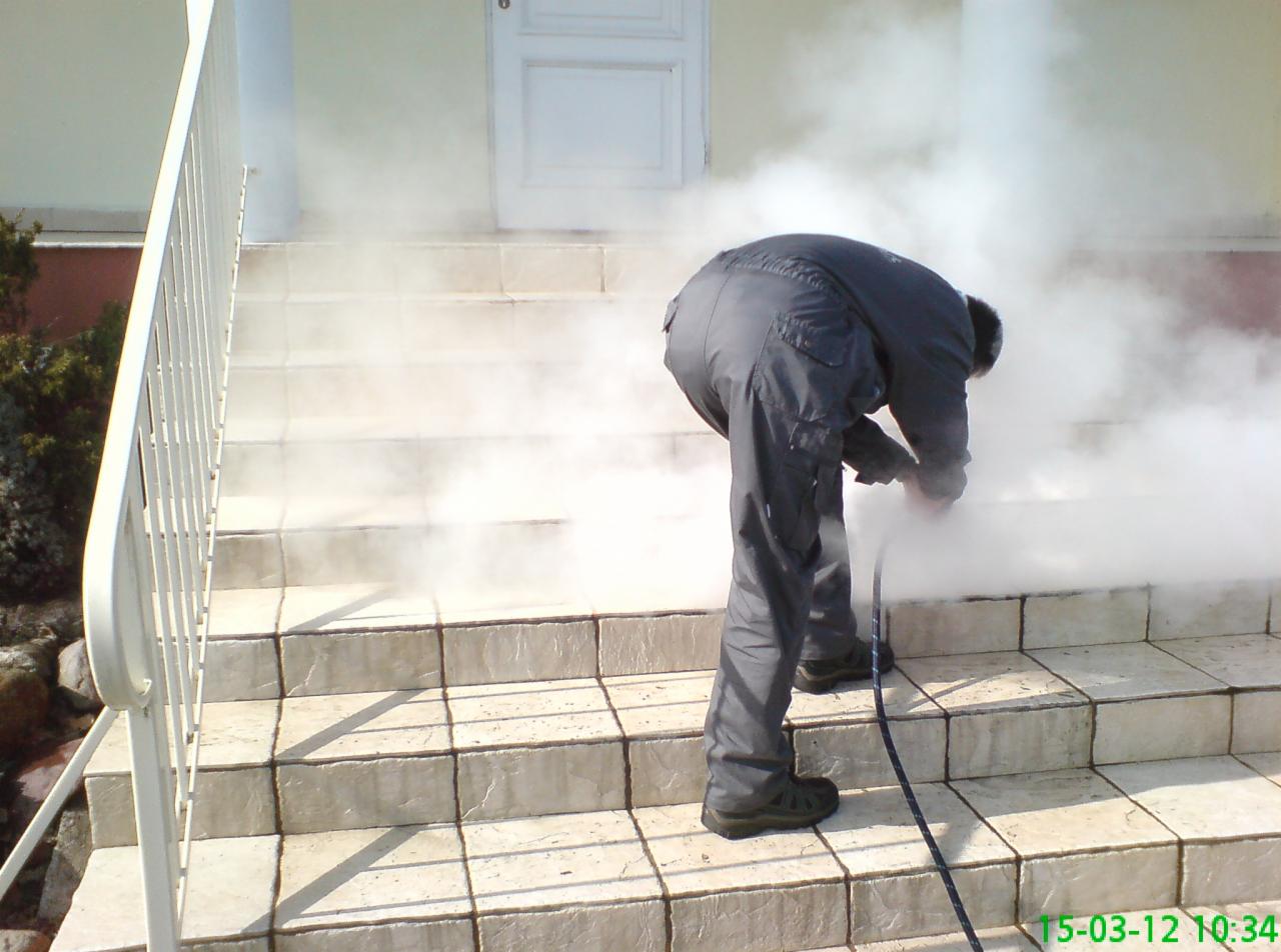 Mężczyzna w ciemnym kombinezonie czyści schody wejściowe do domu za pomocą myjki ciśnieniowej, generując obłoki pary.
