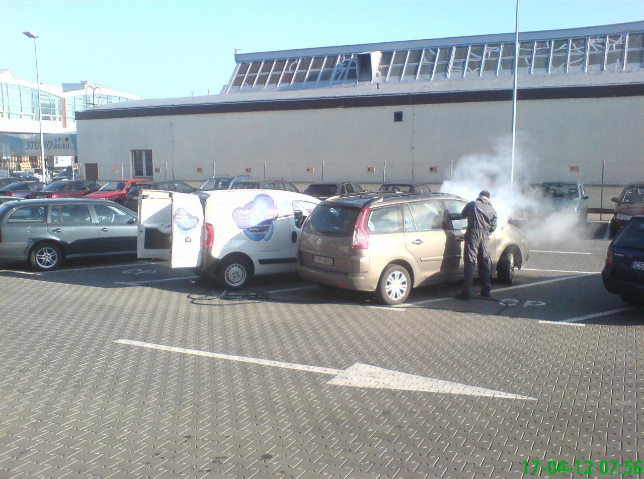 Pracownik w kombinezonie czyści parą wnętrze samochodu na parkingu, obok zaparkowany biały bus z logo firmy.