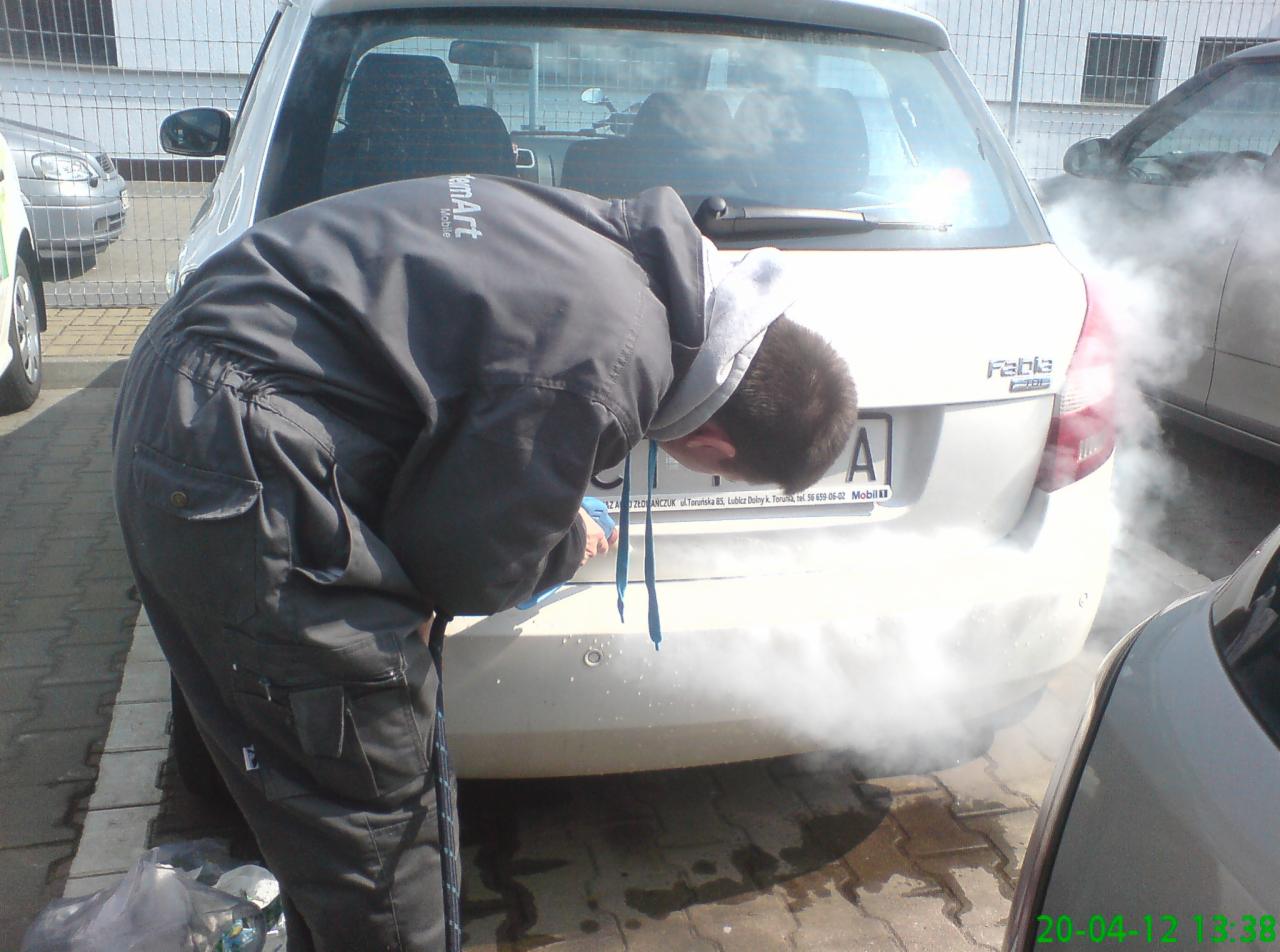 Pracownik w kombinezonie czyści parą tylną część białego samochodu Skoda Fabia, widoczna tablica rejestracyjna i para unosząca się znad mokrej kostki brukowej.