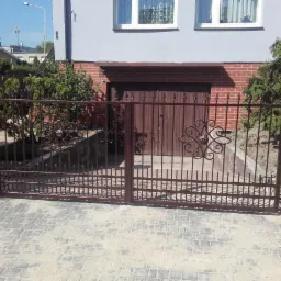 Otwarta, brązowa brama wjazdowa z kutymi elementami dekoracyjnymi, prowadząca na posesję w Starej Dąbrowie, widoczna na tle garażu z brązowymi drzwiami.