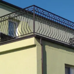 Kuta balustrada balkonowa z dekoracyjnymi elementami na tle zielonej elewacji budynku i błękitnego nieba.