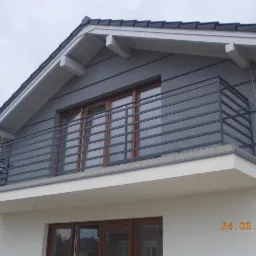 Nowoczesna, pozioma, metalowa balustrada balkonowa w kolorze grafitowym, zamontowana na białym balkonie domu z szarą elewacją, widoczna data 24.06.2013.