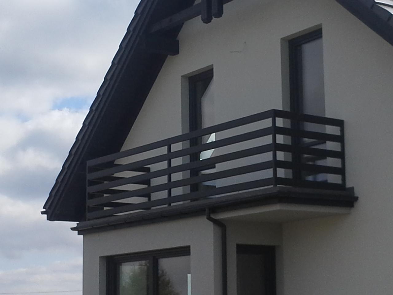 Nowoczesny balkon z czarną, poziomą balustradą na tle szarej elewacji domu jednorodzinnego z widocznym fragmentem dachu i okien.