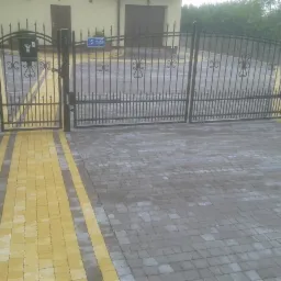Brama wjazdowa z metaloplastyki i kostka brukowa w odcieniach szarości z żółtymi pasami dla osób niepełnosprawnych. Widok z bliska na gotową realizację.