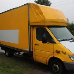 Żółto-biały samochód dostawczy Mercedes-Benz Sprinter z wysoką zabudową, zaparkowany na mokrej nawierzchni, z widoczną białą powierzchnią reklamową na boku.