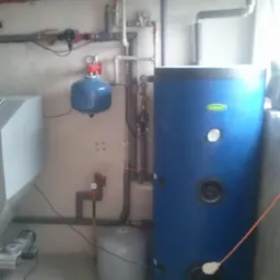 Niebieski zasobnik ciepłej wody użytkowej z widocznymi rurami i zaworami. Szara ściana i podłoga. Elementy instalacji grzewczej w piwnicy.