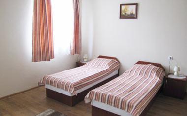 Pokój dwuosobowy z dwoma pojedynczymi łóżkami w kwaterze noclegowej. Na każdym łóżku pościel w biało-brązowe pasy. Przy każdym łóżku stolik nocny z lampką. W tle okno z zasłonami w odcieniach brązu...