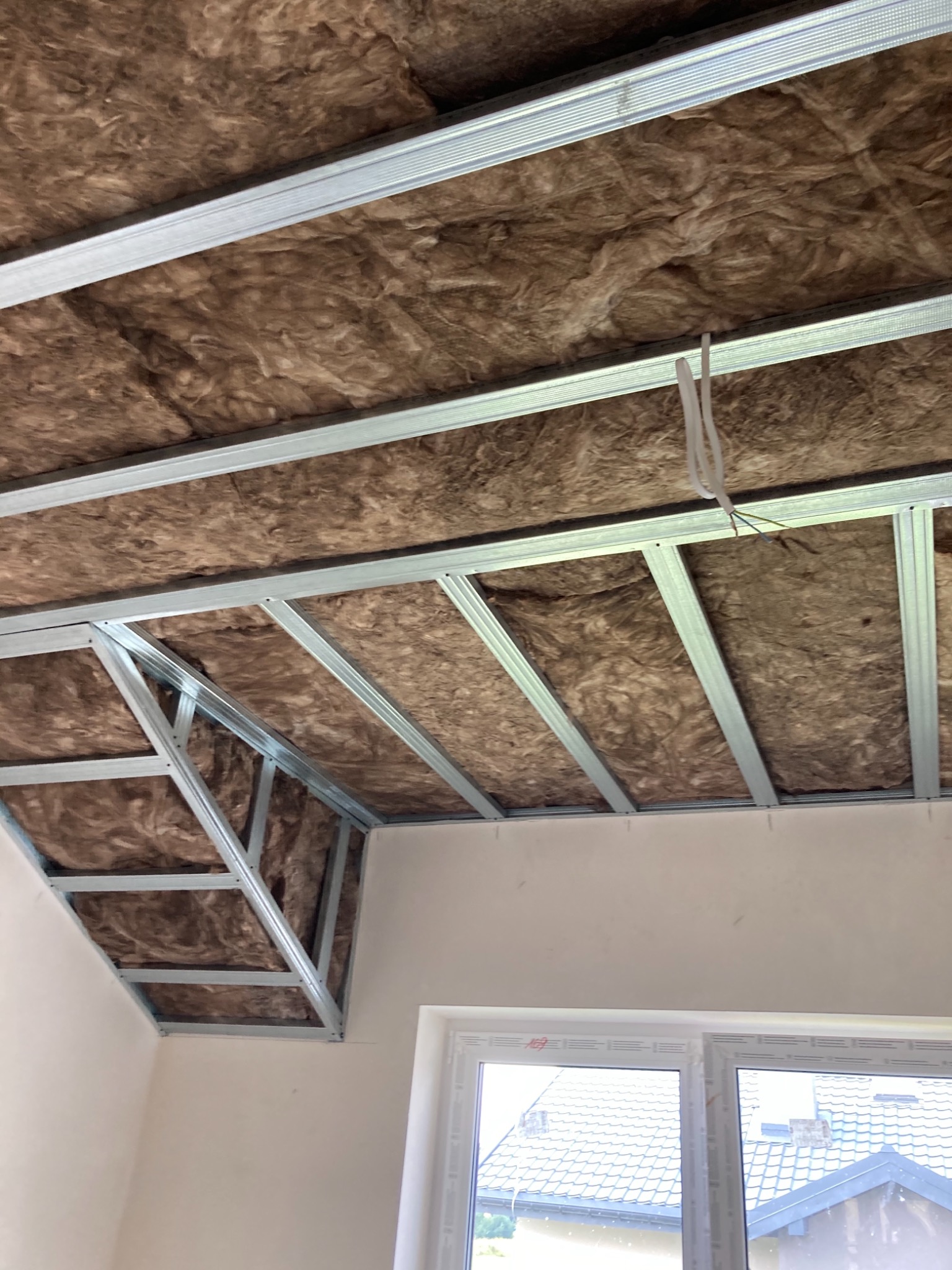 Poddasze w trakcie ocieplania: widoczna wełna mineralna między metalowymi profilami konstrukcji sufitu podwieszanego, wystające kable elektryczne, okno dachowe w tle.