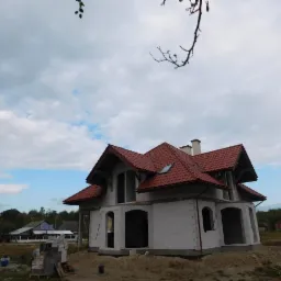 Niedokończony dom jednorodzinny z czerwonym dachem, widoczny z zewnątrz w pochmurny dzień. Budynek w stanie surowym, z widocznymi pustakami i otworami okiennymi.