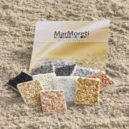 Prezentacja próbek dekoracyjnych kamieni w kwadratowych formach na tle piaszczystej powierzchni, z widocznym katalogiem marki Marmoreti Natural Colors.