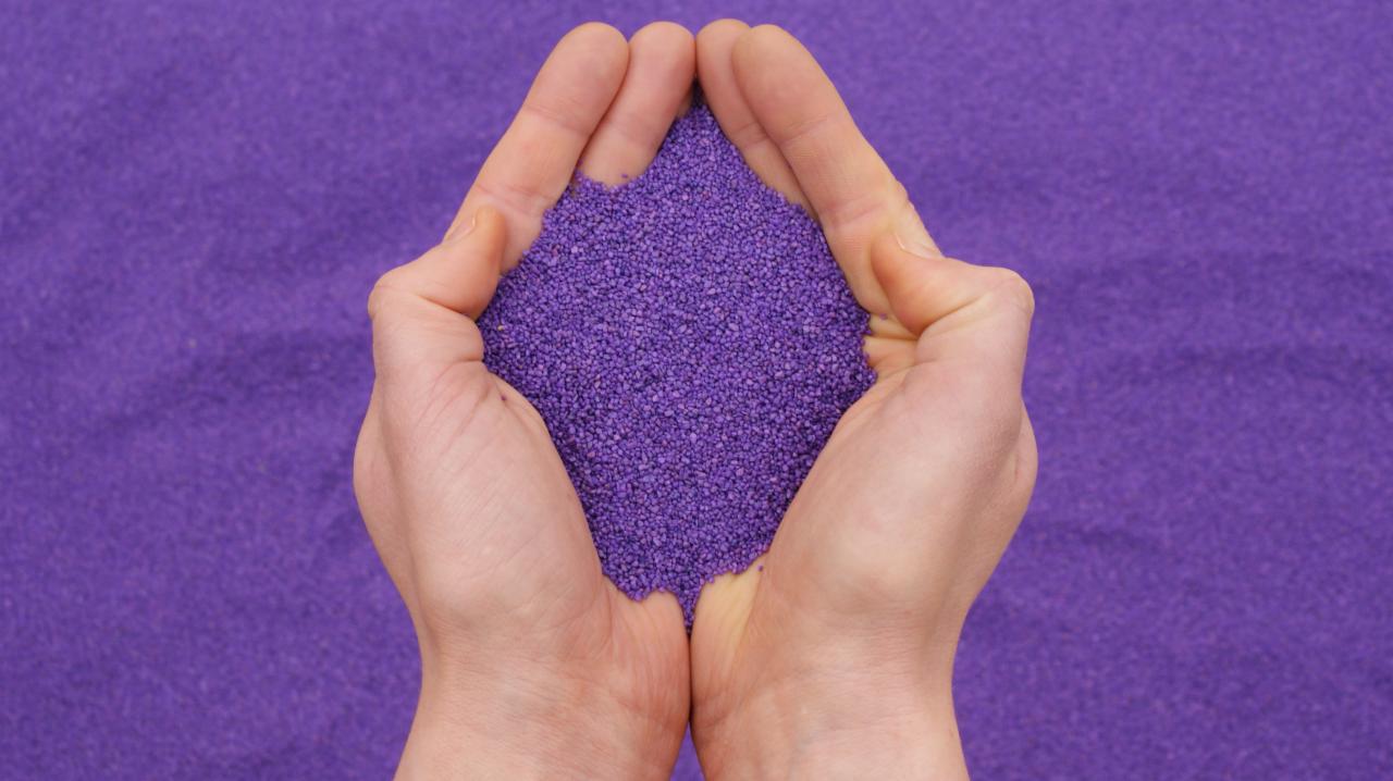 Dłonie trzymające garść drobnego, fioletowego kruszywa na tle fioletowego podłoża.