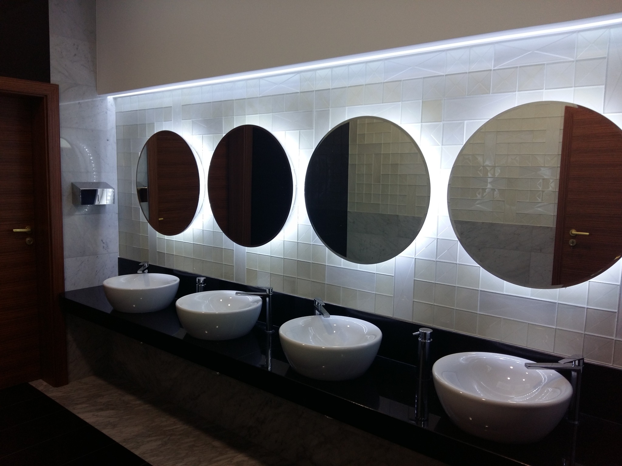 Nowoczesna łazienka publiczna z czterema okrągłymi lustrami z podświetleniem LED, białymi umywalkami nablatowymi i czarnym blatem.