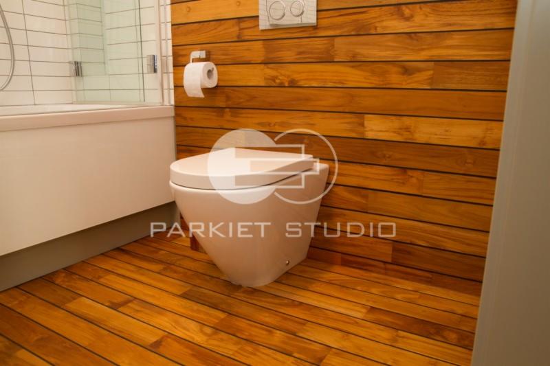 Nowoczesna łazienka z drewnianą podłogą i ścianą z paneli, wisząca toaleta, biała wanna z szybą, drewno w ciepłym odcieniu brązu.